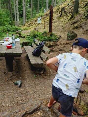 Kind steht an einem Holztisch mit Bänken am Goldbrunnen im Wald, daneben eine Kraxe