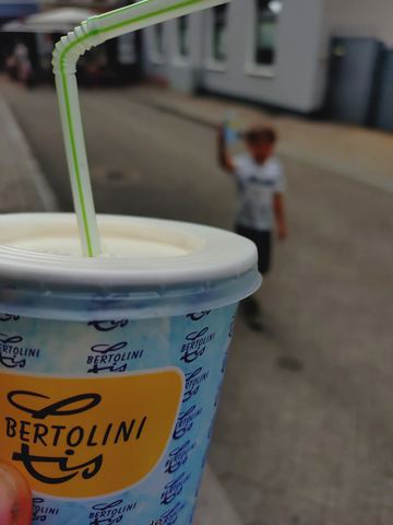 Becher Eis mit Trinkhalm im Vordergrund, dahinter unscharf ein Kind auf einer Straße