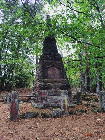 Pyramide aus rotem Sandstein im Wald, umgeben von niedrigen Steinen und einer Bank