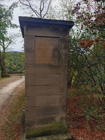 Schlichter Sandsteinpfeiler mit Tafel für Johann Wolfgang von Goethe am Rand eines Waldwegs, daneben eine Bank