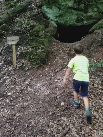 Kind läuft auf einen Felsvorsprung mit dunkler Höhlenöffnung zu, davor ein Hinweisschild