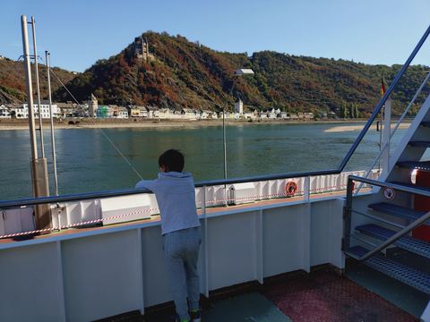 Kind steht von hinten fotografiert an der Reling einer Fähre und schaut auf den Rhein und die Häuser am gegenüberliegenden Ufer
