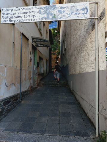 Verwittertes Schild über einer schmalen Gasse mit der Aufschrift „Fußweg zum Stadtteil Heide, Hallenbad, Hühnertempel, Freilichtbühne, Loreley über Burg Katz (keine Besichtigung)“