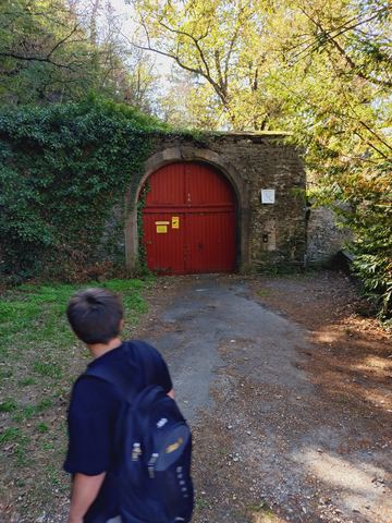 Rotes Rundbogentor in einer efeubewachsenen Bruchsteinmauer im Wald, davor ein Kind mit Rucksack