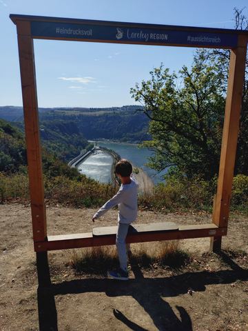Kind balanciert von hinten fotografiert auf der Querlatte eines großen hölzernen Bilderrahmens, durch den man auf den Rhein sieht