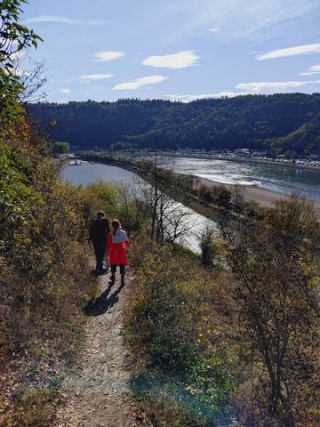 Zwei Personen gehen von hinten fotografiert einen schmalen, steinigen Pfad steil bergab, darunter der Rhein mit einer Sandbank