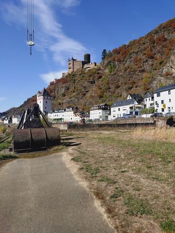 Blick vom Rheinufer hinauf zu Burg Katz auf einem herbstlich verfärbten Hang, im Vordergrund ein alter Hafenkran